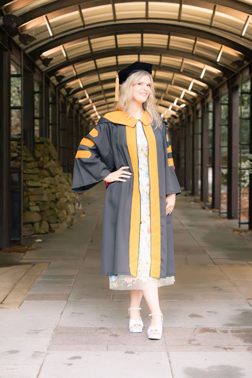 Smiling graduate in dark academic gown with gold trim and cap poses in a glass-roofed walkway.