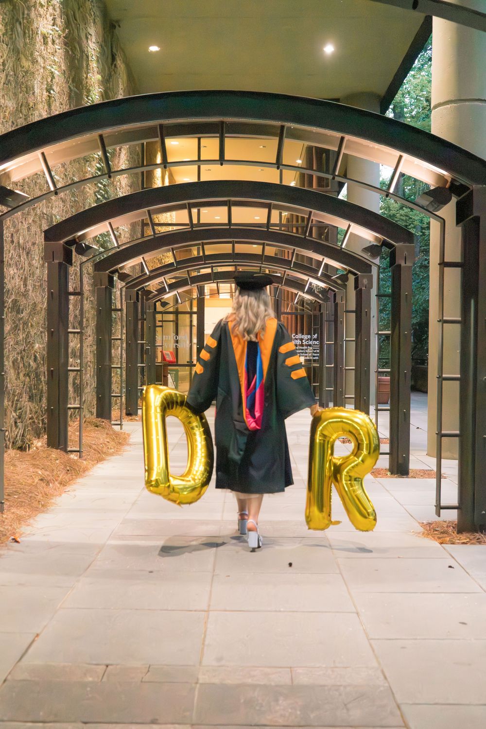 Graduate in cap and gown holds gold "DR" balloons while walking through an arched outdoor corridor.