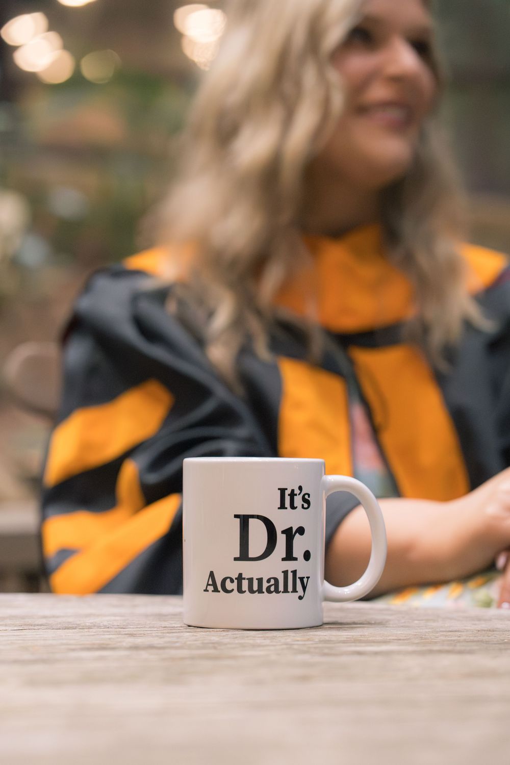 White mug reading Its Dr. Actually on wooden table, blurred woman in graduation regalia smiling in background.