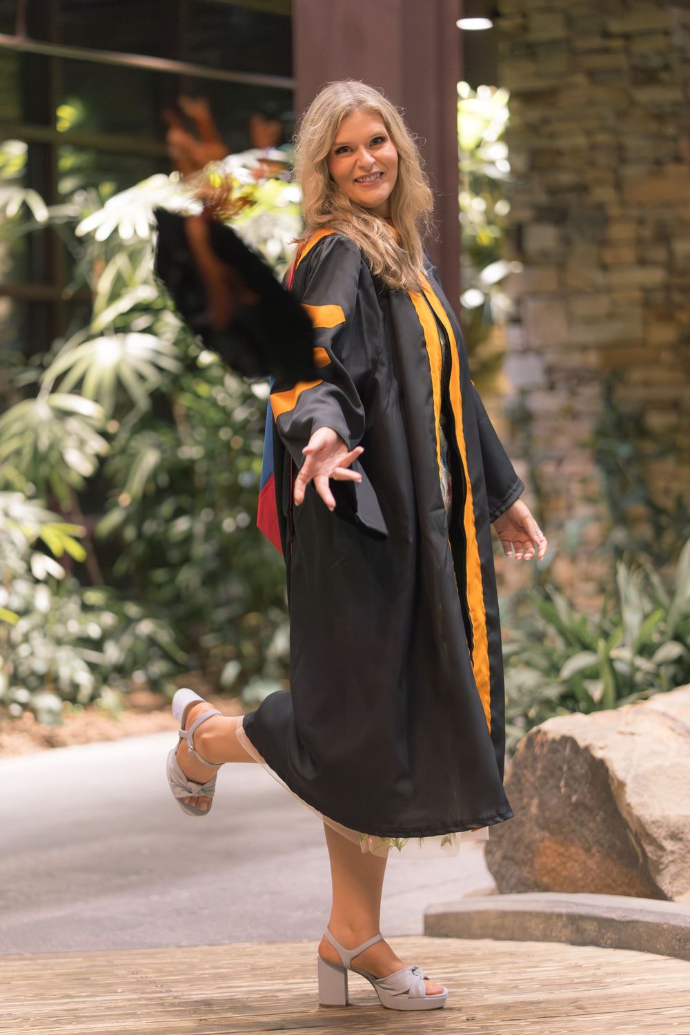 A joyful woman twirls in a black graduation gown with gold trim, celebrating outdoors among lush greenery.