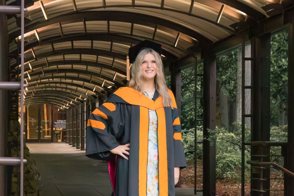 Woman in doctoral graduation regalia with orange hood poses in an arched glass corridor surrounded by trees.