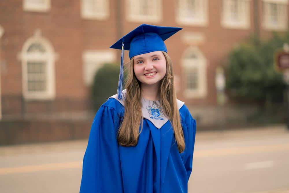 Graduate in blue regalia smiling and posing against brick building.