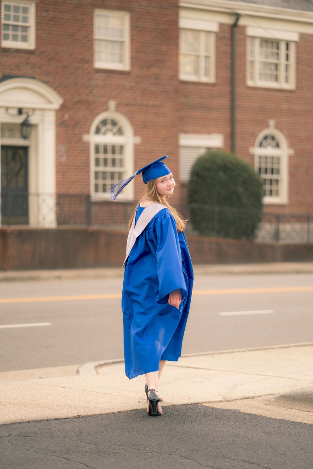Graduate in blue cap and gown posing playfully on campus sidewalk.