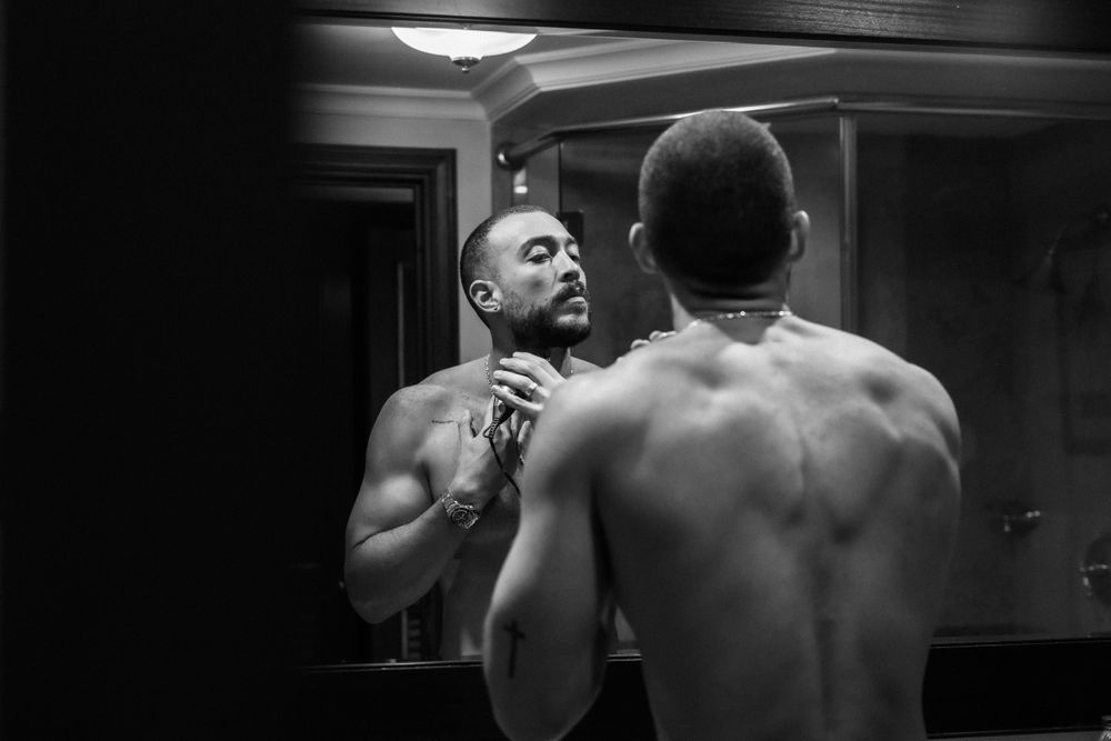 Man shaving his beard while looking in bathroom mirror, black and white artistic portrait showing muscular back.