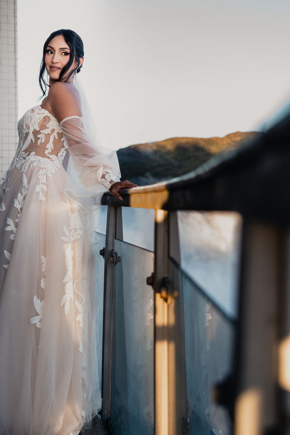 Bride in flowing lace gown poses elegantly on balcony during golden hour.