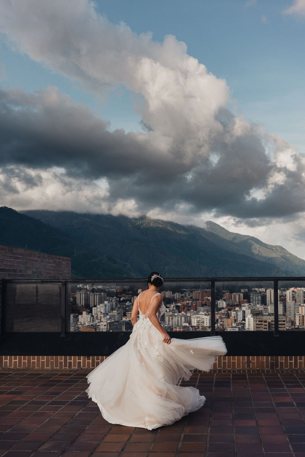 A bride in a flowing white dress twirls on a rooftop terrace overlooking a cityscape with mountains and dramatic clouds.