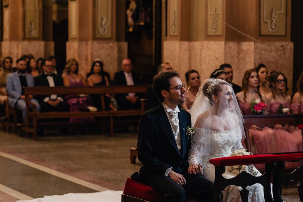 Couple seated during traditional wedding ceremony in ornate church with wooden pews and decorative details.