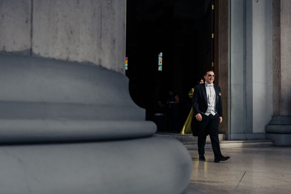 A person in formal attire stands near grand architectural columns in dramatic lighting.