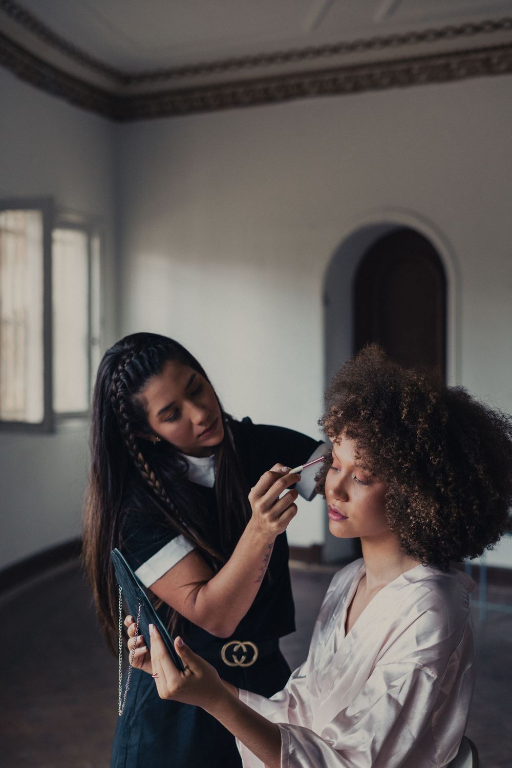 Makeup artist applying cosmetics on a client while checking phone reference in an elegant interior setting.