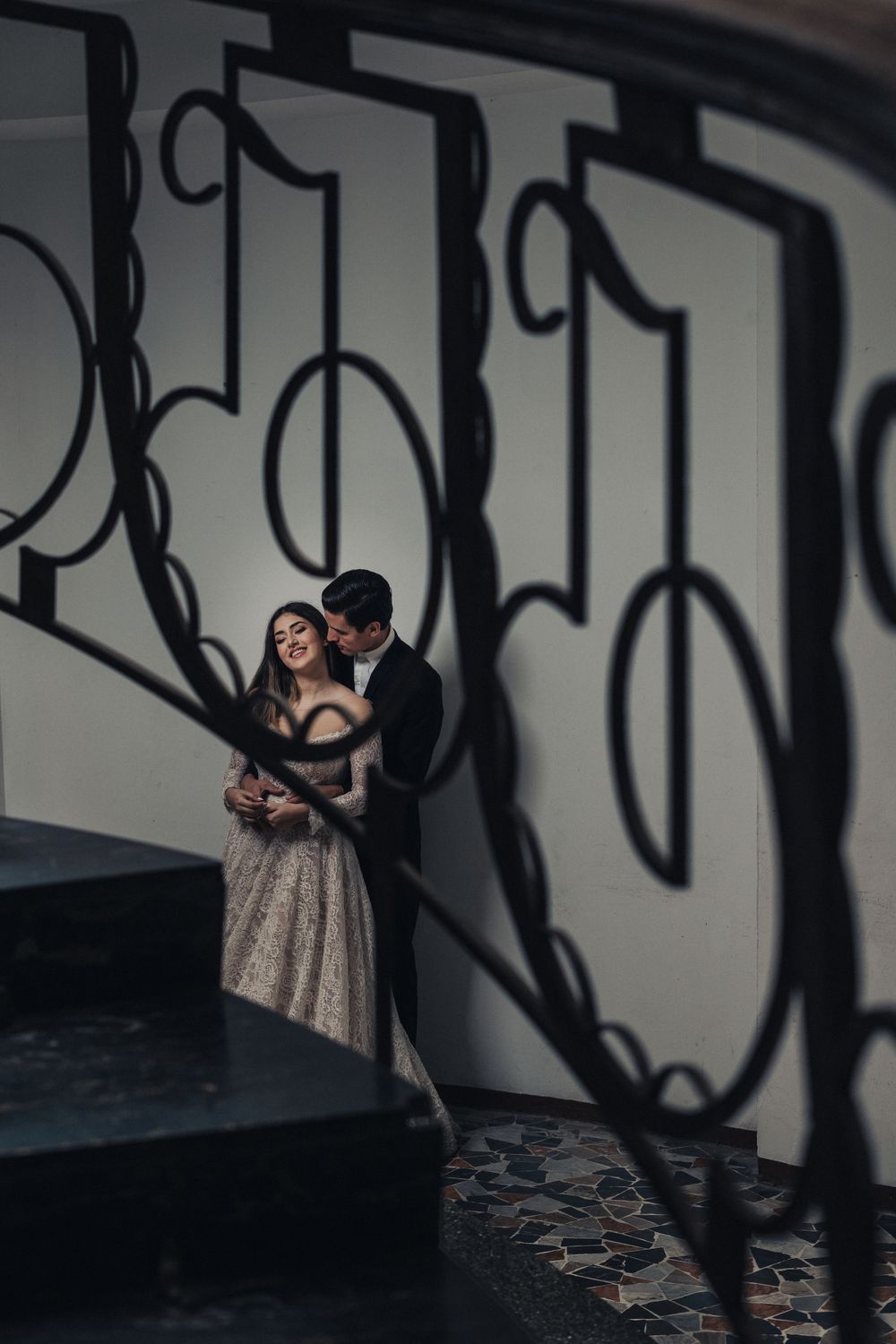A romantic couple dances in a dimly lit space with decorative ironwork shadows cast on the wall behind them.