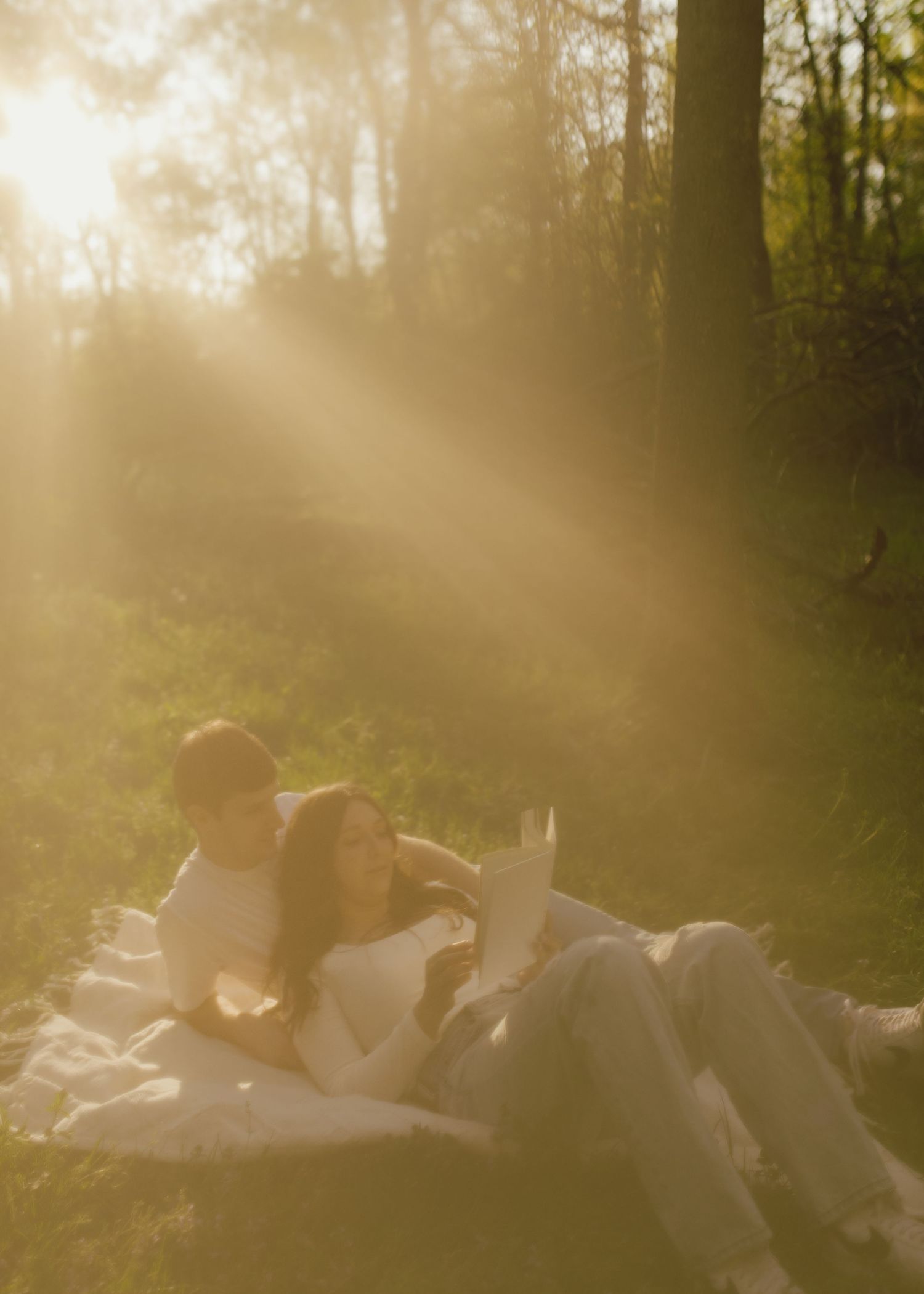 Silhouetted couple shares tender moment in sunlit field as person in white dress stands with partner kneeling at sunset.