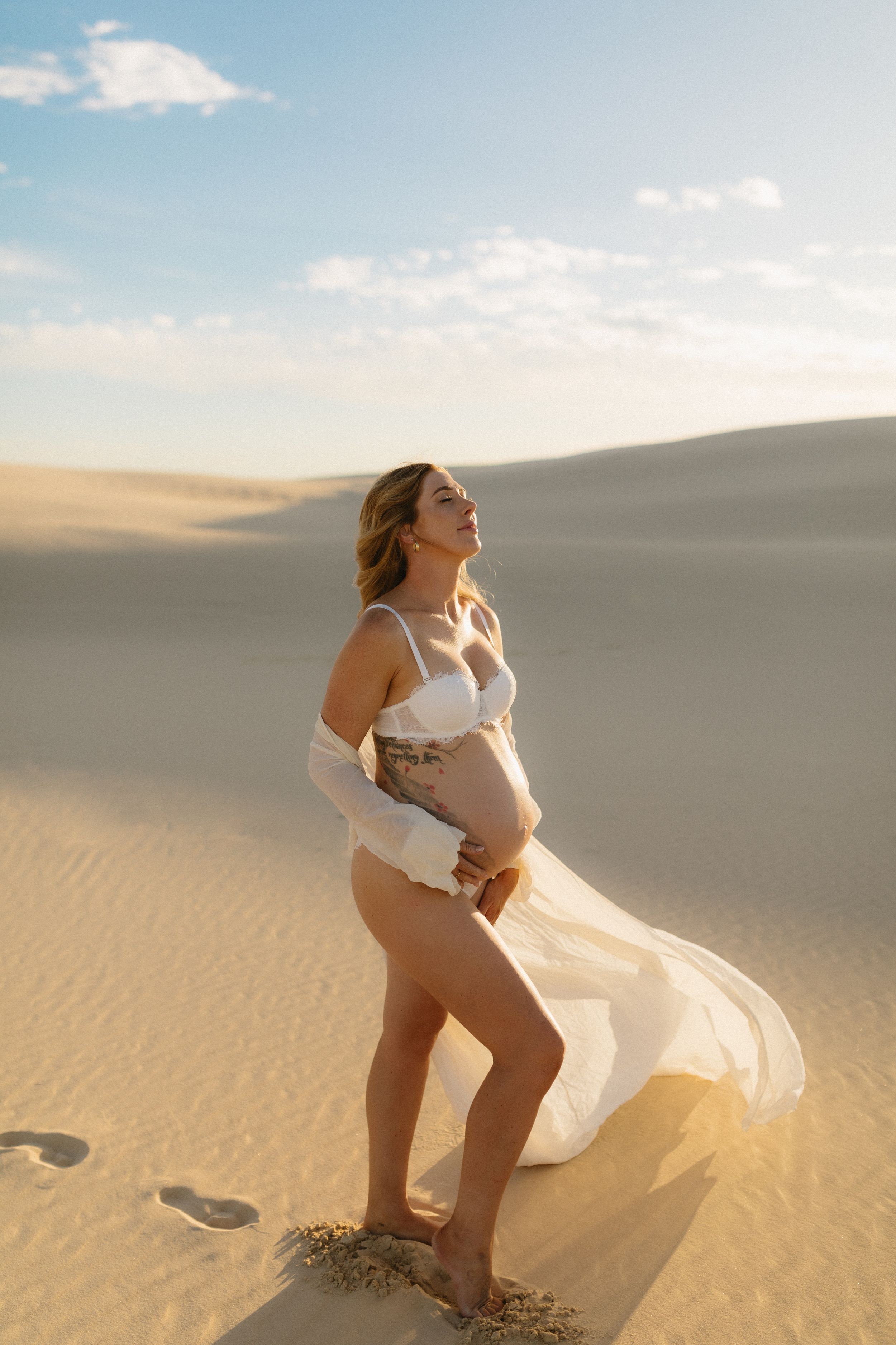 Expectant mother in flowing white dress poses gracefully on desert sand dune.