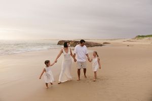 Family of four in white outfits walking hand-in-hand on a sandy beach at sunset.