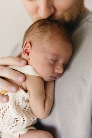 A father tenderly kisses his sleeping newborn baby wrapped in a cream knit outfit.