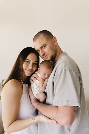 Young couple smiling and holding their sleeping newborn baby wrapped in lace, posing for a family portrait.