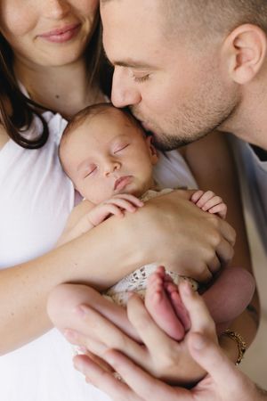 Parents lovingly holding and kissing their sleeping newborn baby, captured in a tender family portrait.
