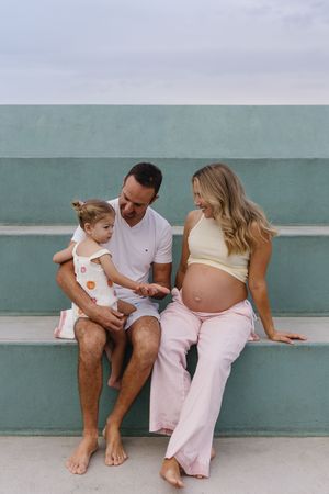 Pregnant woman in yellow crop top and pink pants sits with partner and toddler on outdoor steps near teal wall.