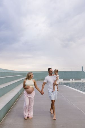 Pregnant woman holding hands with man carrying toddler, walking along waterfront promenade under cloudy sky.