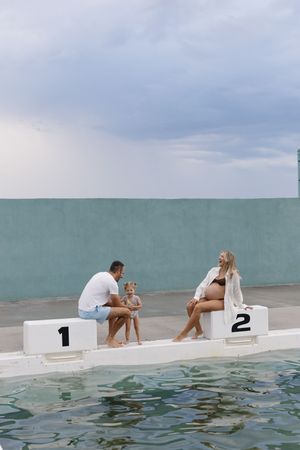 Pregnant woman and man with child sitting on numbered podium blocks beside a pool with cloudy sky backdrop.