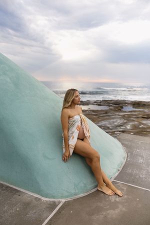 Pregnant woman wrapped in floral fabric leaning against teal sculpture by rocky ocean shore at sunset.