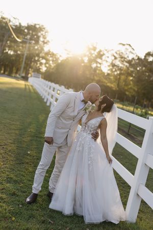 Bride and groom sharing a romantic kiss at golden hour beside a white fence on a lush green lawn.