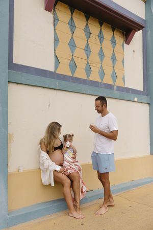 Pregnant woman sitting with toddler beside man in blue shorts, posing by colorful vintage beach building.