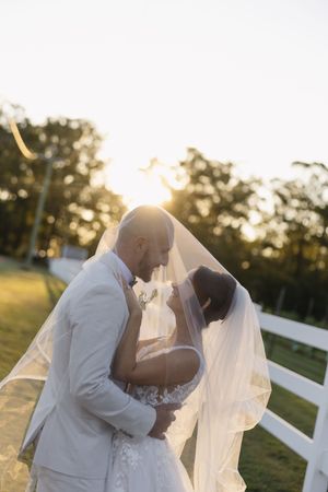 Bride and groom share romantic moment under veil at golden hour, white fence and trees in background.