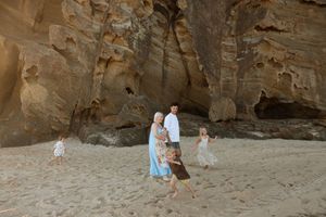 Family walking on sandy beach near large rock cave formations, children playing around parents in natural coastal setting.