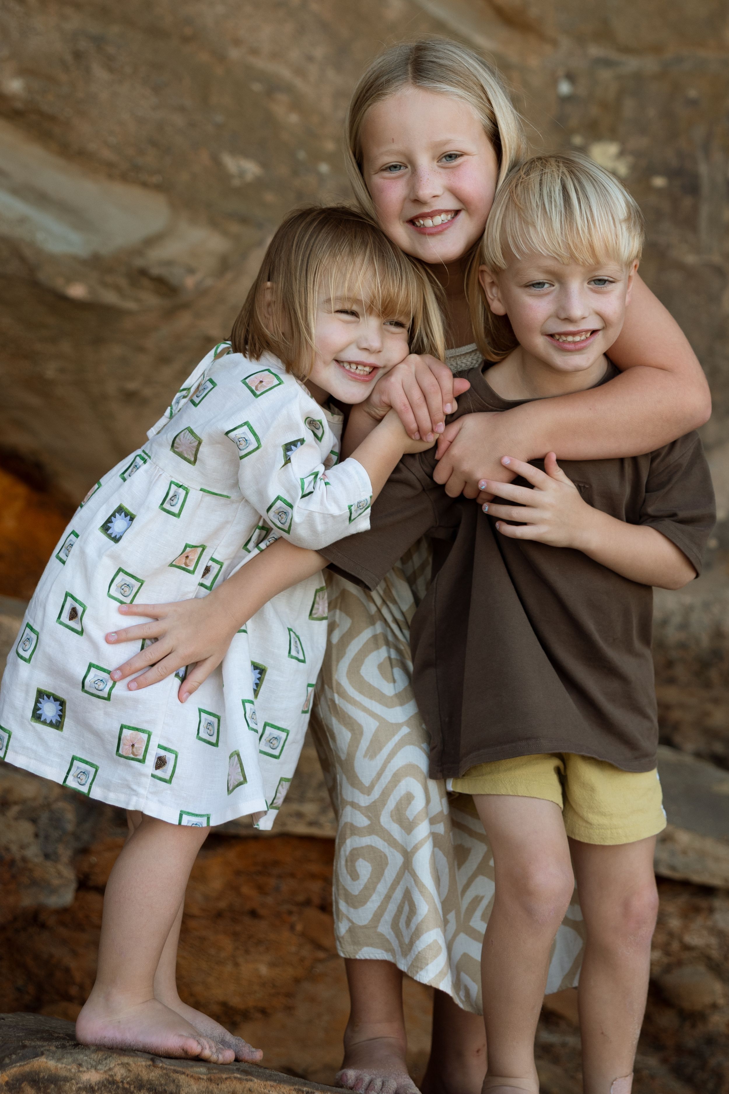 Three smiling blonde children hugging each other outdoors on rocks, wearing casual summer clothing.
