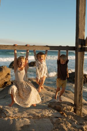 Three happy children hanging from a wooden beam at a sandy beach with ocean waves in the background.