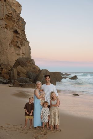 Family of five posing on a sandy beach at sunset with rocky cliffs and ocean waves in the background.