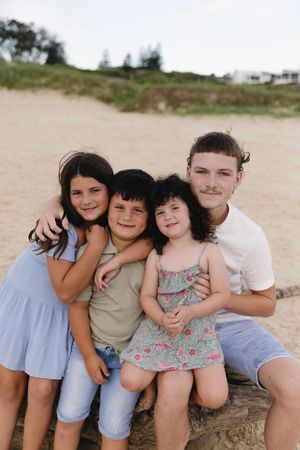 Four siblings smile together on a sandy beach, with the oldest teen boy hugging the younger children.