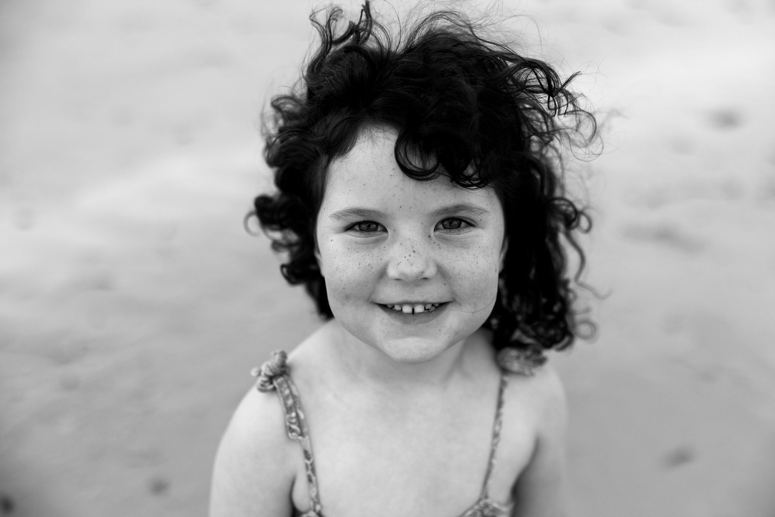 Black and white portrait of a smiling curly-haired little girl with freckles at the beach.