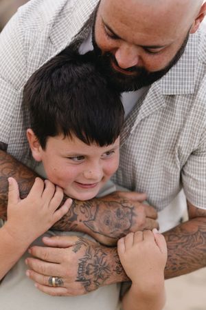 Tattooed father with wedding ring hugs young son from behind, sharing a tender moment in casual outdoor setting.