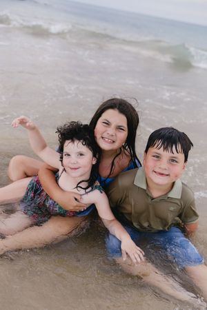 Three happy children playing together in ocean waves on a sandy beach, smiling and posing for a photo.