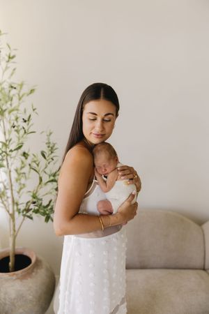 A young mother in a white dress tenderly holds her sleeping newborn baby close to her chest indoors.