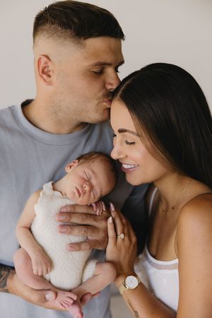 A couple tenderly holds their sleeping newborn baby, with the father kissing the mother's forehead in a studio portrait.