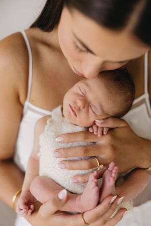 A mother tenderly kisses her sleeping newborn baby wrapped in a soft white blanket.
