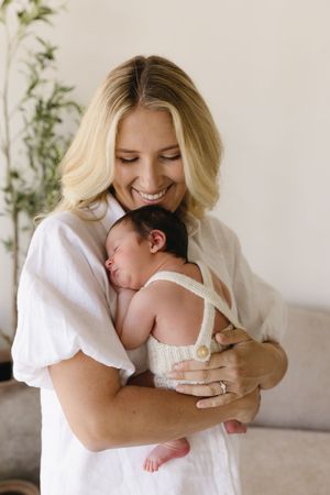 Smiling blonde mother in white dress tenderly holding her sleeping newborn baby against her chest.