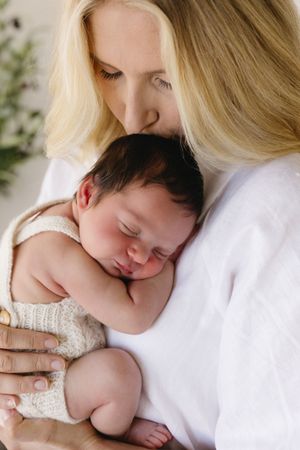A blonde woman tenderly kisses a sleeping newborn baby wrapped in a cream knit blanket.
