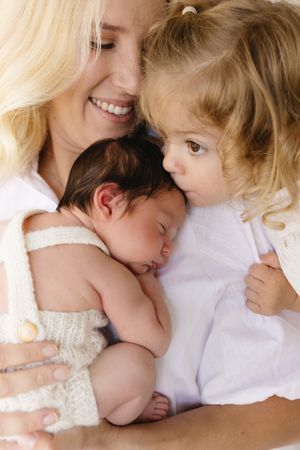 Smiling blonde mother holds sleeping newborn baby while young toddler girl leans in to give a gentle kiss.