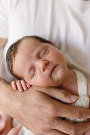 A newborn baby sleeping peacefully in an adult's hands, wearing a white knit outfit.