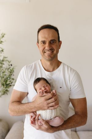 Smiling father in white Tommy Hilfiger t-shirt proudly holds his sleeping newborn baby wrapped in a cream knit blanket.
