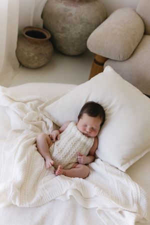 Newborn baby sleeping peacefully on white pillow and knit blanket, wearing cream romper, with rustic pottery in background.