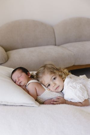 A toddler girl with blonde curls lies beside a sleeping newborn baby on a white bed near a gray sofa.