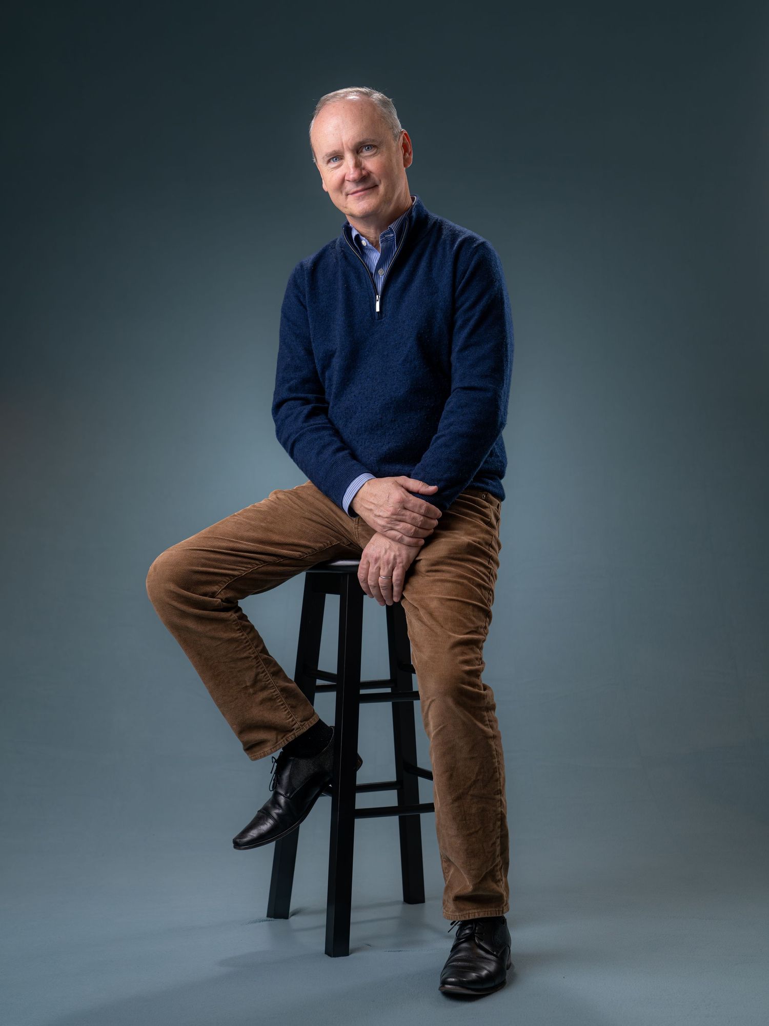Man in navy sweater and tan pants sits on a black stool, smiling, against a gray studio background.