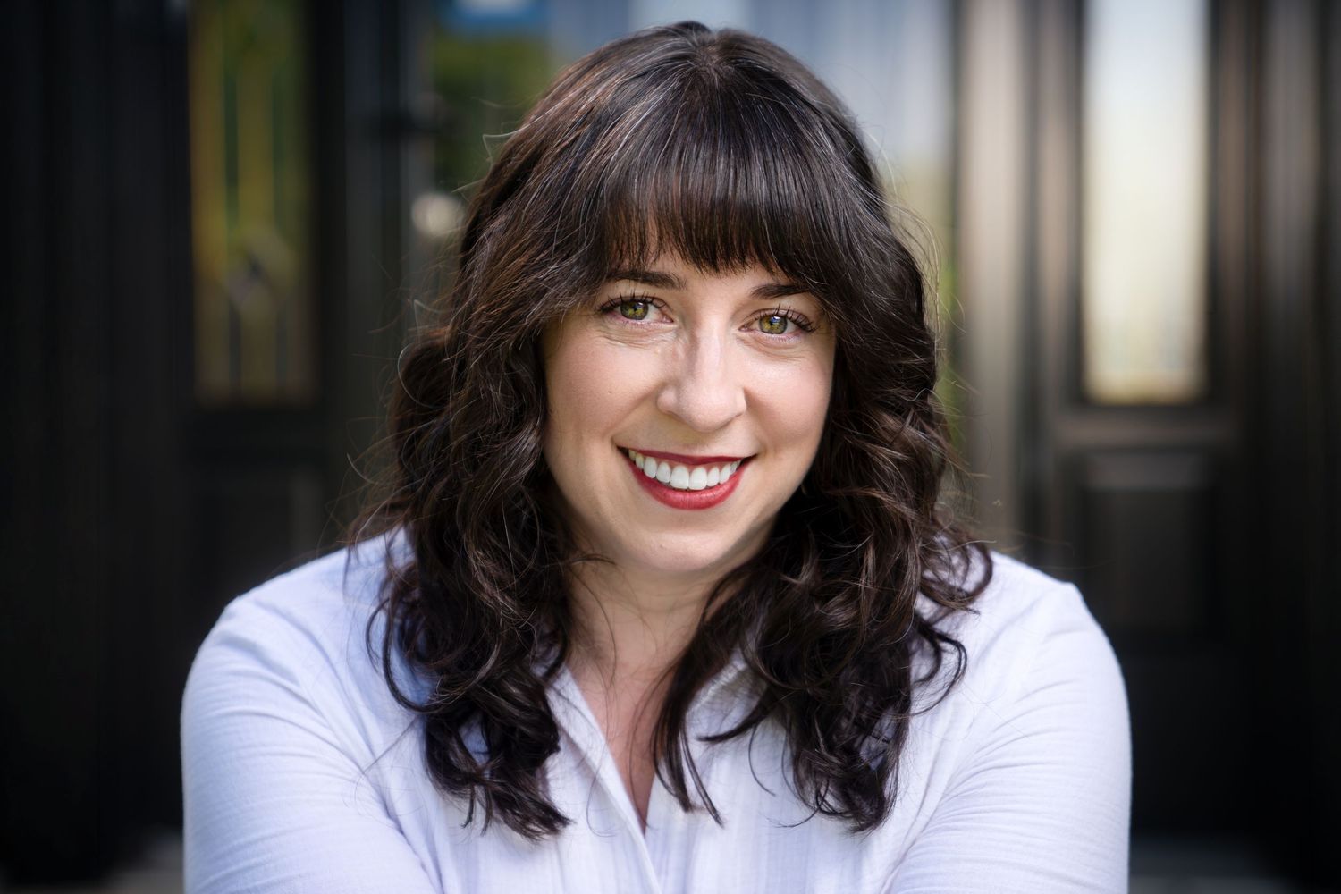 Smiling woman with dark curly hair poses outdoors in white shirt for professional headshot.