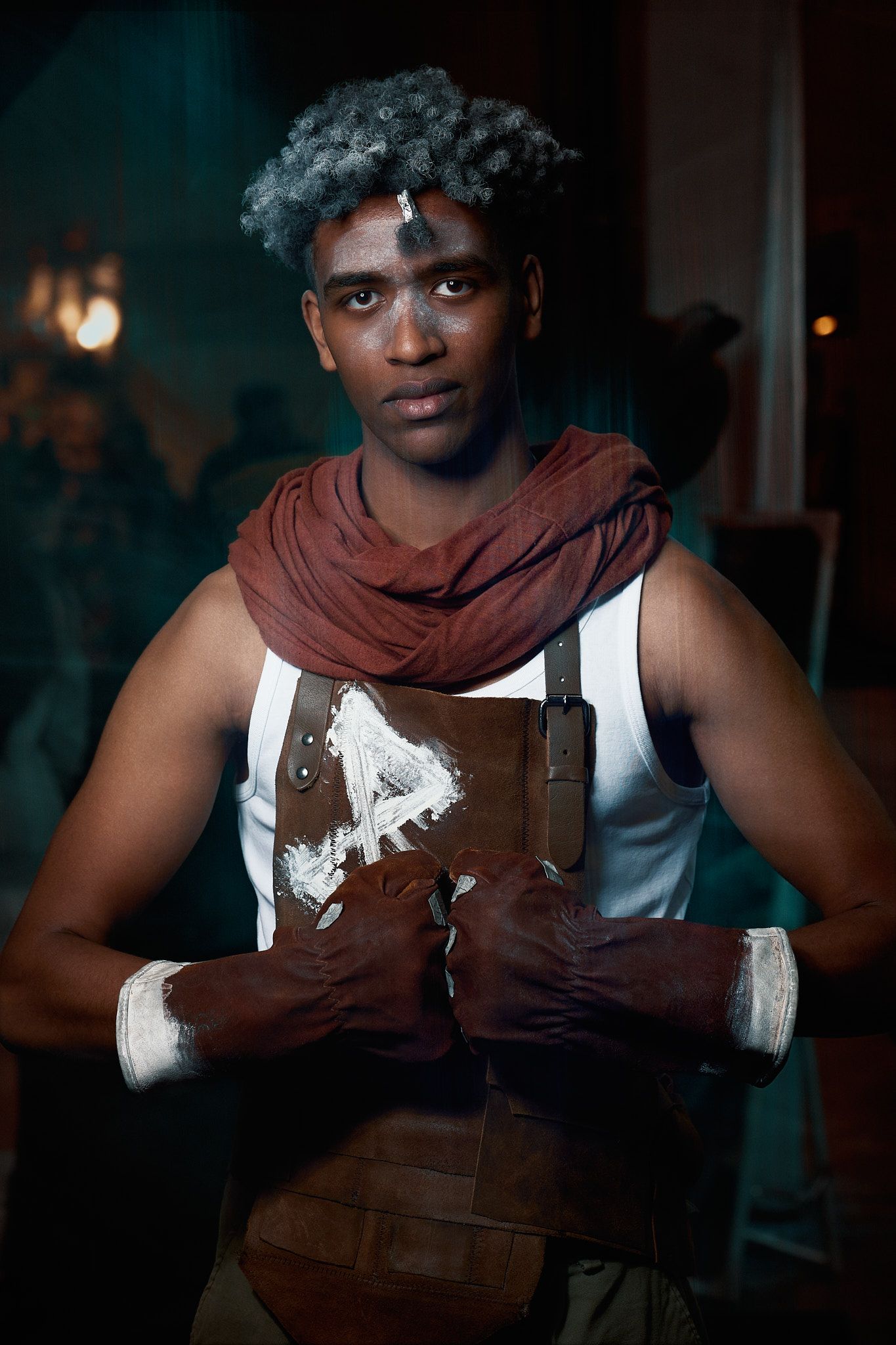 A person wearing white and brown overalls with a maroon scarf against a dark background poses in dramatic lighting.