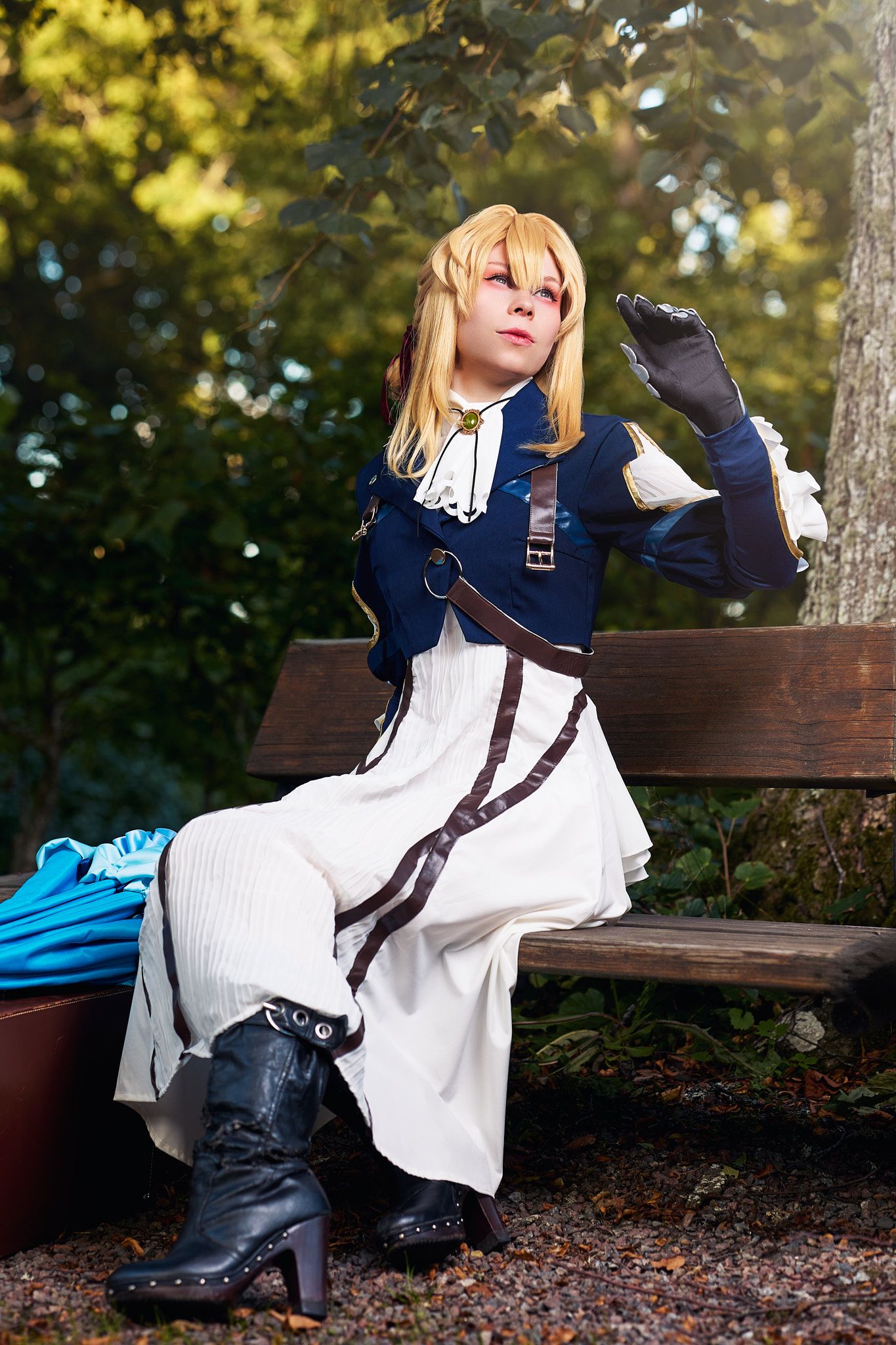 A cosplayer in a navy and white dress poses dramatically on a wooden bench in an outdoor autumn setting.