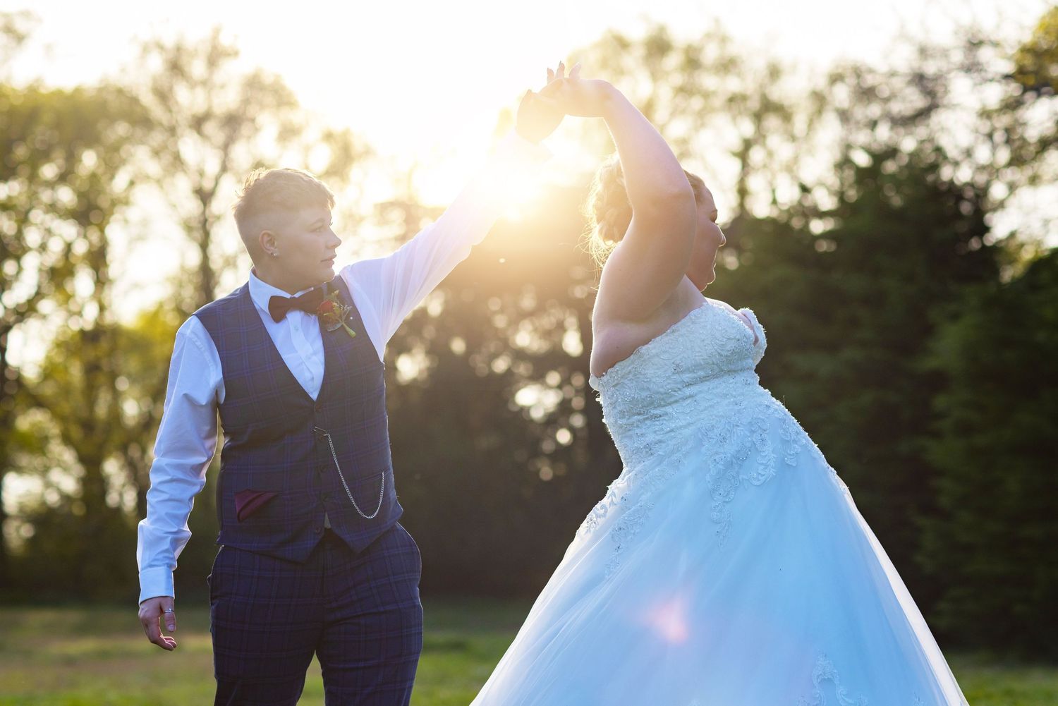 Same-sex couple dancing outdoors at sunset, with one partner in a white wedding gown and the other in a vest and shirt